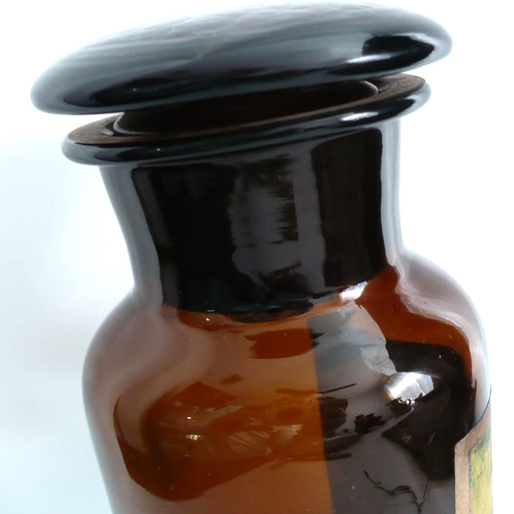 19th-century hand-blown amber glass apothecary jar with a label indicating it once contained cardamon seed powder. The jar has a dark amber color, a narrow neck, and a round stopper.