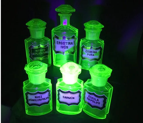 A collection of antique uranium glass apothecary jars and bottles glowing bright green under UV blacklight