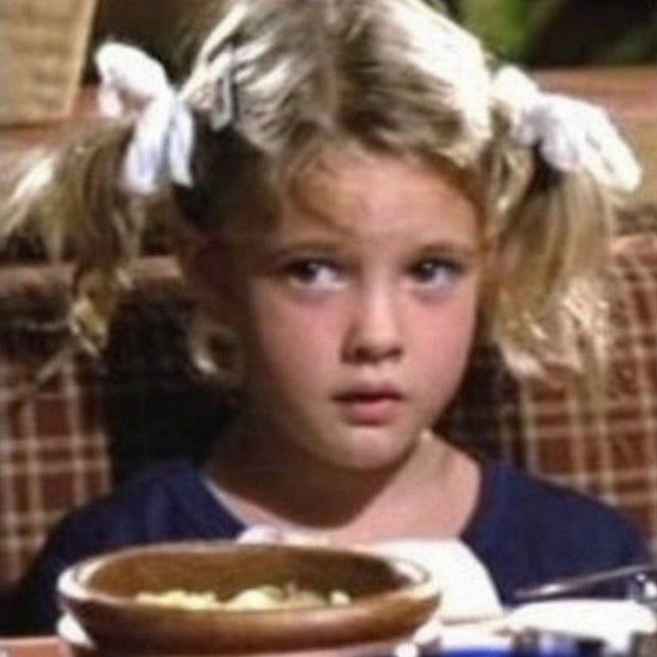 A young girl with blonde pigtails tied with white yarn hair bows, wearing a navy blue top, seated at a table with a bowl of food in front of her. Gertie costume.