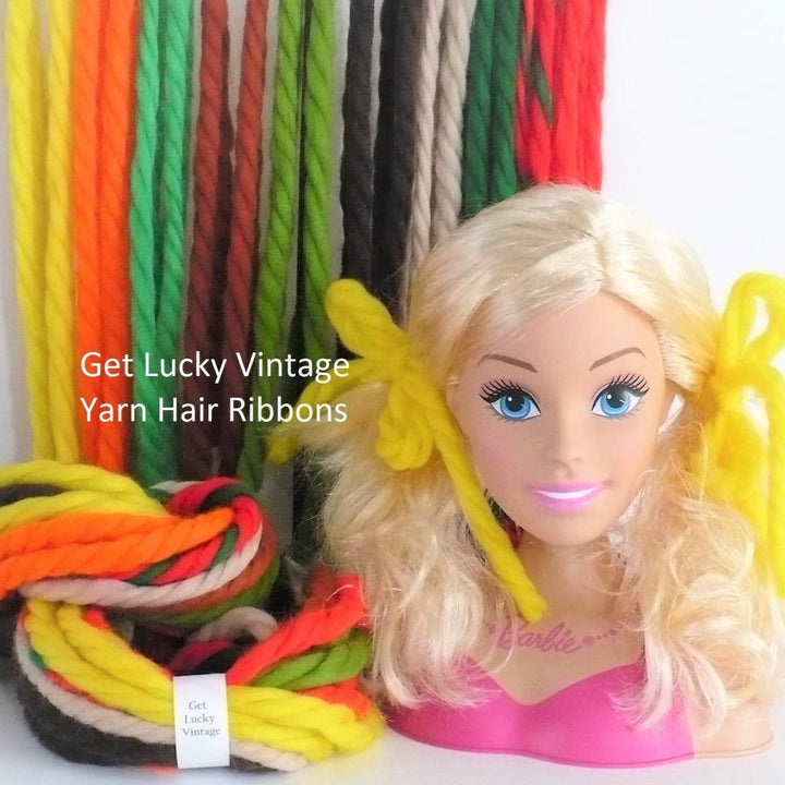 A blonde Barbie doll bust wearing a pink top, styled with bright yellow yarn hair ribbons in two pigtails, set against a display of many hanging ribbons and a bundle of colorful yarn ribbons.