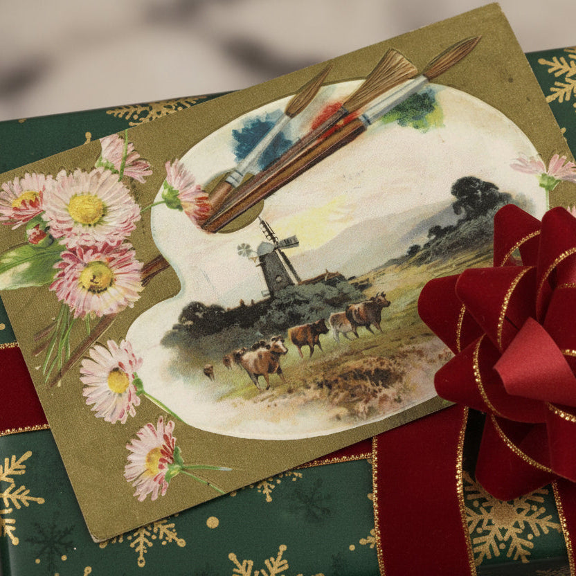Gift box with a 1907 Painted Tidings From An Artist’s Palette Vintage Christmas Postcard and red ribbon