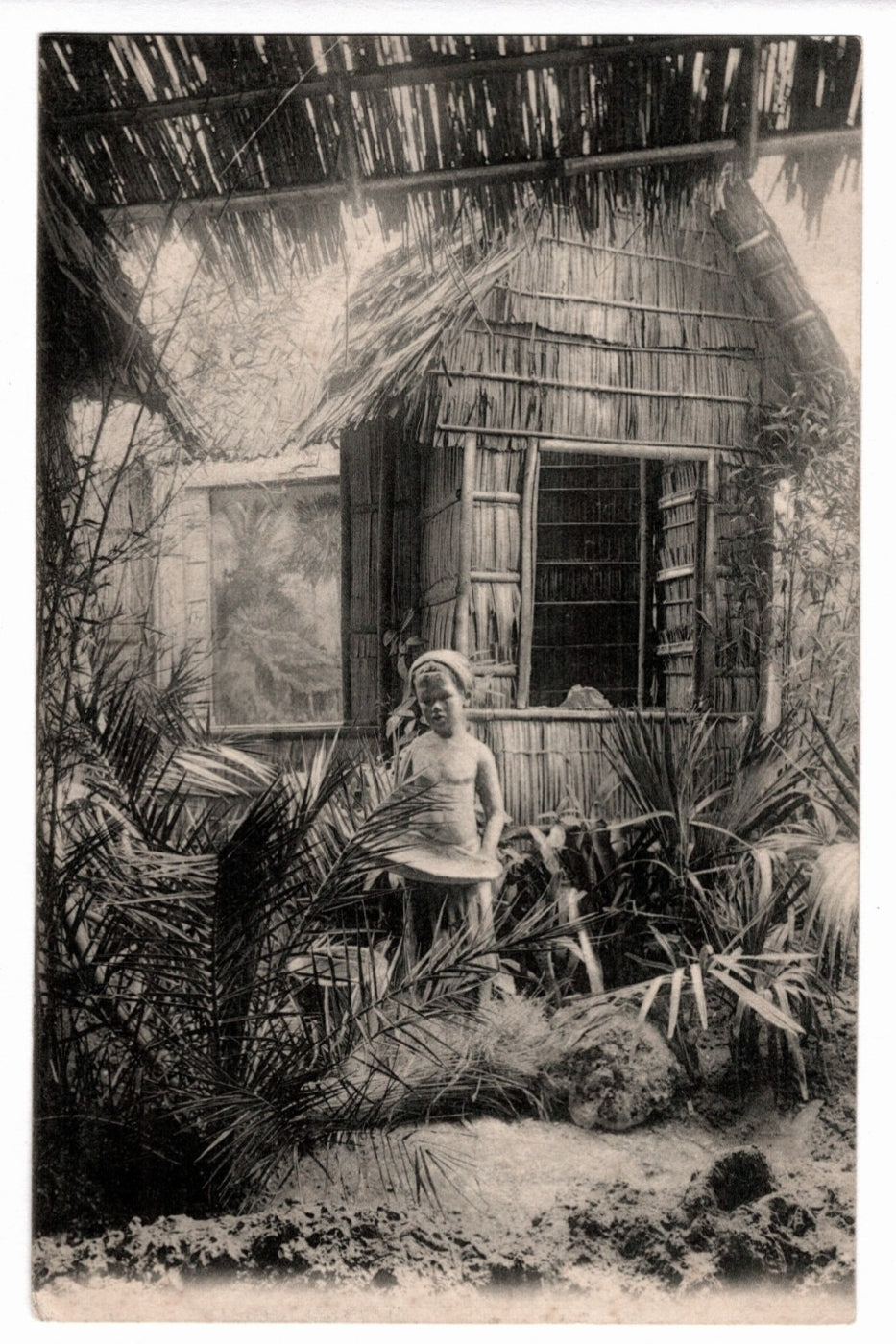  A young child standing before a traditional rural dwelling constructed with thatched palm fronds and bamboo. The lush tropical setting includes various palm plants and a shaded, open-sided structure in the foreground. 