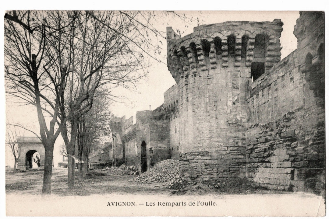 This antique monochromatic postcard depicts a striking section of the medieval fortifications in Avignon, France, known as Les Remparts de l’Oulle. The image captures the weathered stone towers and curtain walls that surround the historic center of the city. In the background, the Porte de l’Oulle (Oulle Gate) is visible. The foreground shows a rustic, unpaved path with bare trees, highlighting the historic atmosphere of the city before modern urban development.