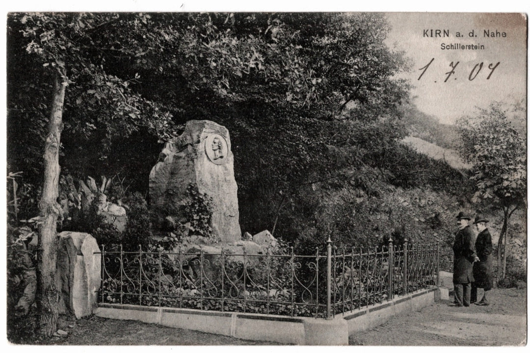 Vintage postcard of a stone monument with a coat of arms, surrounded by trees and people in a park-like setting. the monument consists of a large unhewn stone featuring a profile relief portrait, enclosed by an ornamental wrought-iron fence. Two men in period attire are seen standing next to the memorial, which is nestled in a wooded area.