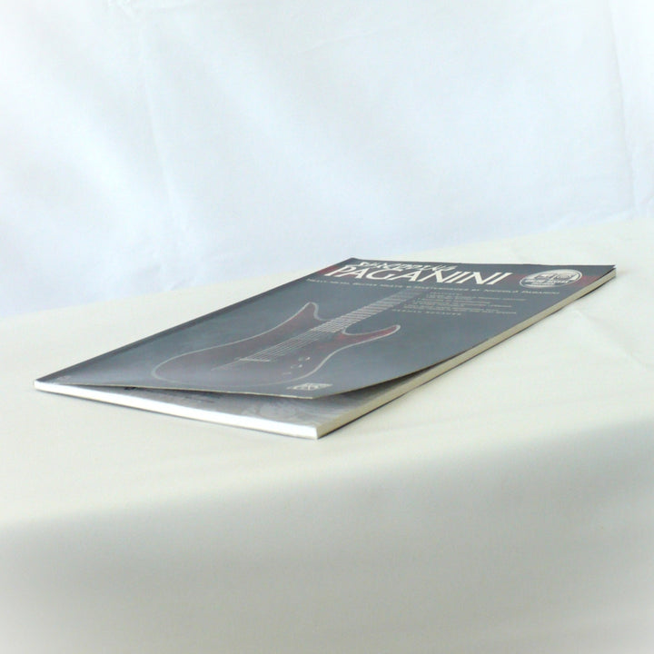 "Lower corner detail of Shredding Paganini guitar tab book showing a gentle natural curve with no hard creases or bends.