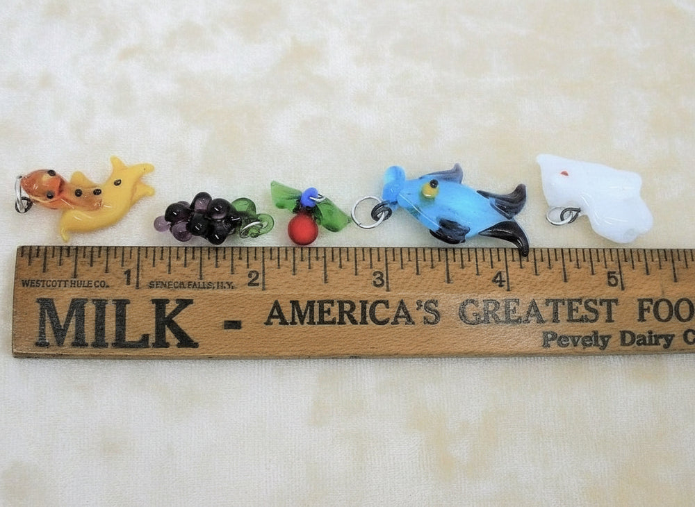 1960s vintage lampwork charms: a white elephant, a salamander, a fruit, a heart, a blue fish, and a grape cluster next to ruler.