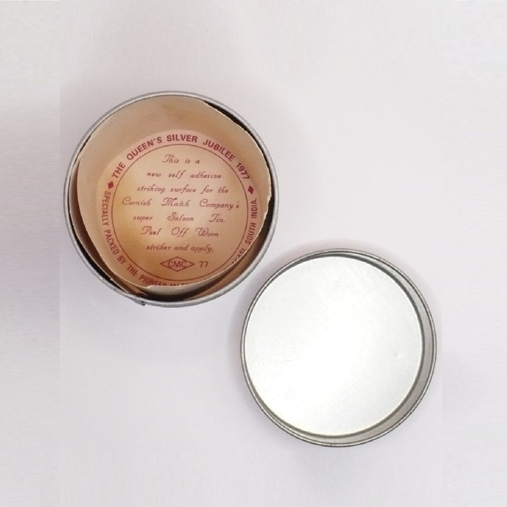 An open round metal Matchbox Tin of silver jubilee tea, showing the light brown tea liquid inside and the silver-colored metal lid resting beside it on a white background.