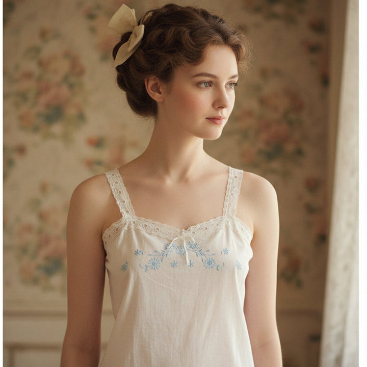 Woman wearing a white embroidered Woman wearing a white Edwardian Cambric Corset Cover in a softly lit room with floral wallpaper.