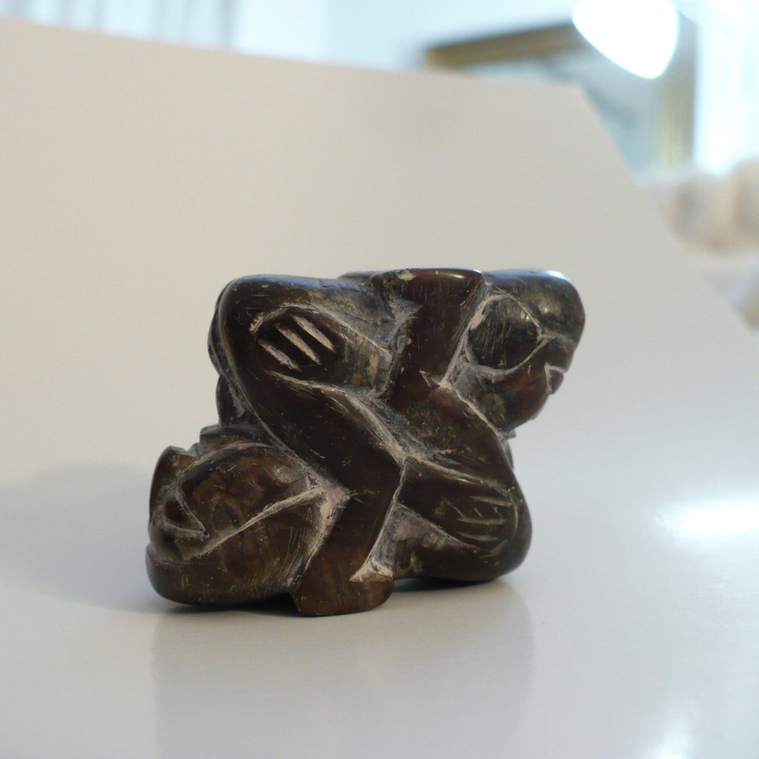 A small carved dark brown stone sculpture depicting two figures embracing, photographed on a light-colored surface with a blurred indoor background. 1970s Erotic Mexican Folk Art Carving.