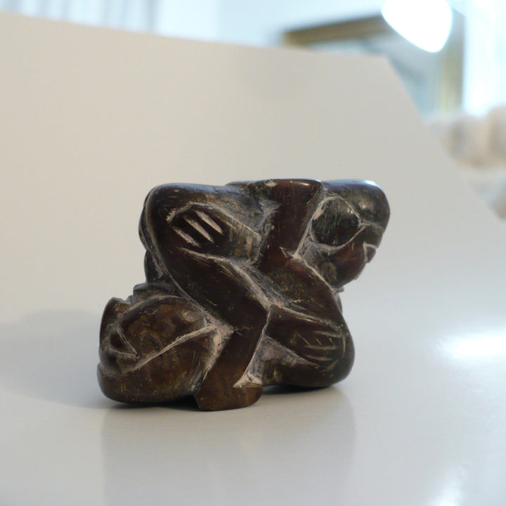 A small carved dark brown stone sculpture depicting two figures embracing, photographed on a light-colored surface with a blurred indoor background. 1970s Erotic Mexican Folk Art Carving.
