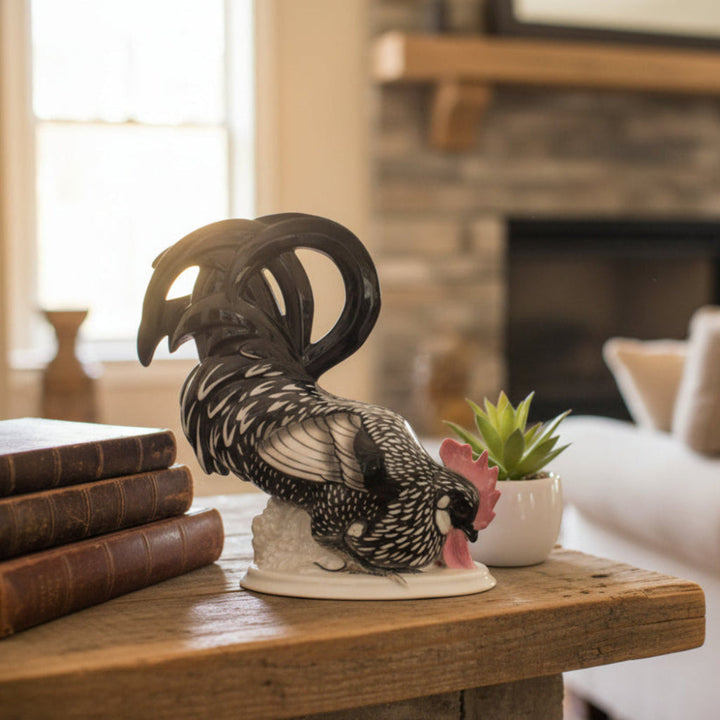 A decorative rooster figurine with black, white, and gray feather detailing and a pink comb, displayed on a rustic wooden table in a cozy living room setting next to a small potted succulent.