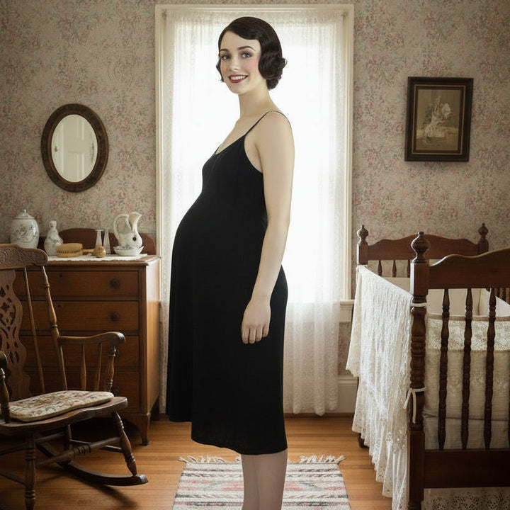Woman in a ntique maternity slip standing in a baby's room with wooden furniture and framed pictures.