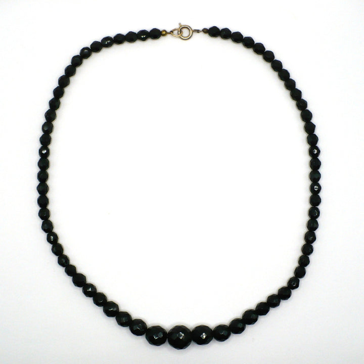 Vintage 1950s single-strand black faceted glass beaded necklace laid out in a circle against a white background.