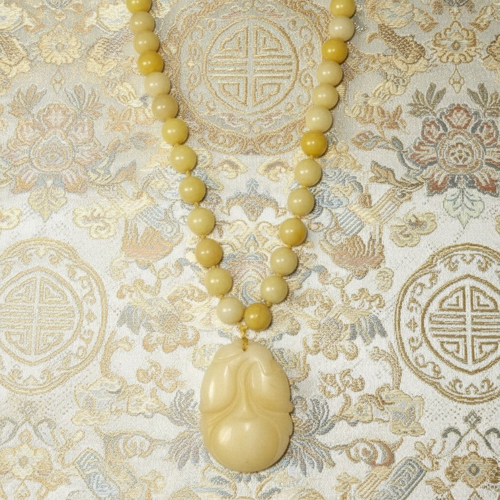 Vintage 1970s yellow nephrite jade beaded necklace featuring a hand-carved persimmon pendant laid flat against an ornate traditional golden silk background.