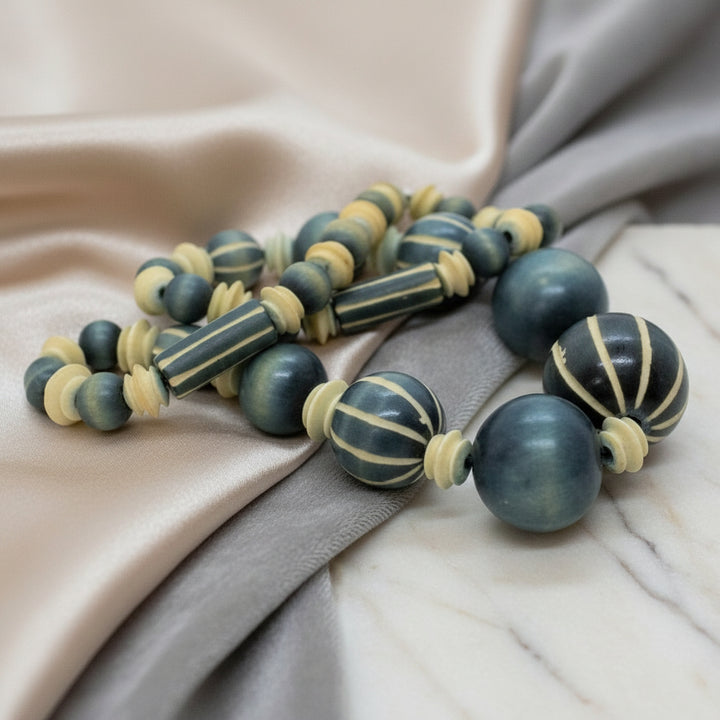1970s vintage beaded necklace featuring blue-grey and cream striped wood beads and carved camel bone spacers resting on fabric.