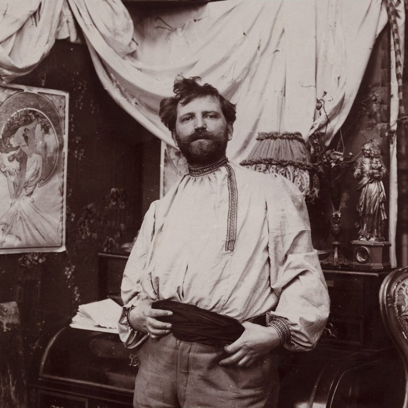 A sepia-toned vintage portrait of a bearded man in a loose, long-sleeved shirt with a dark sash, standing indoors in a decorated room with draped fabric, wall art, and furniture visible.