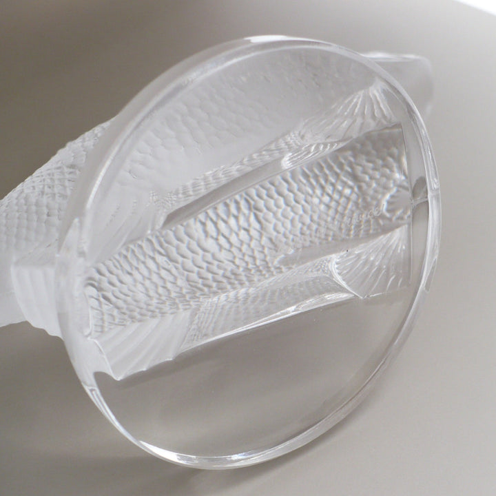 A close-up of a A frosted crystal fish sculpture, Perche Bouchon de Radiateur: Perch Car Mascot, displayed on a clear glass stand against a neutral beige background, base underside.