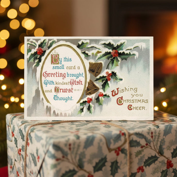 1911 Gilded Bells & Holly Traditional Vintage Christmas Postcard on a wrapped gift with blurred lights in the background