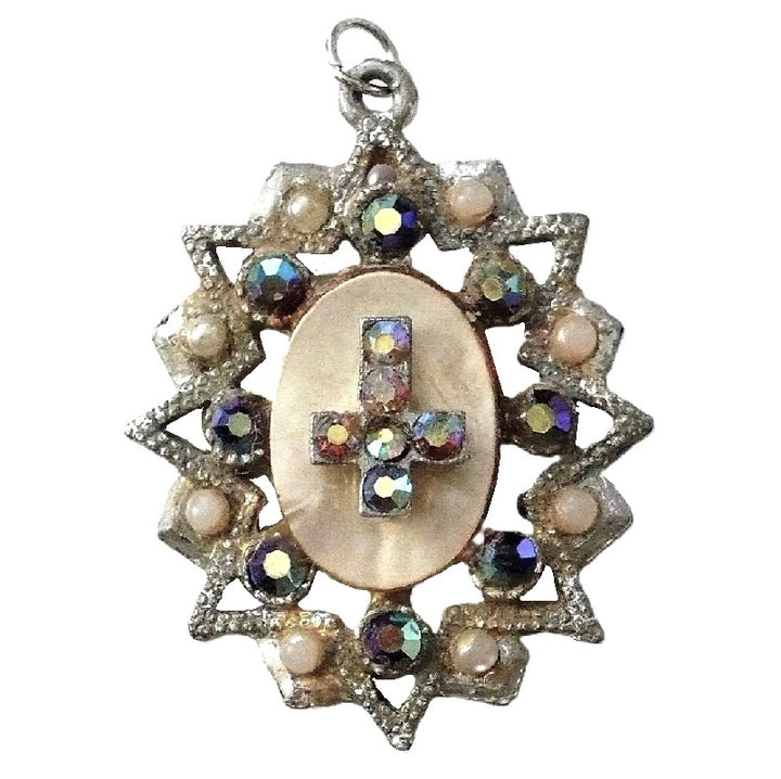 A vintage 1950s pendant featuring a cross with faceted Aurora Borealis stones and simulated pearls, set against an oval mother-of-pearl center, with intricate eloxal metalwork.