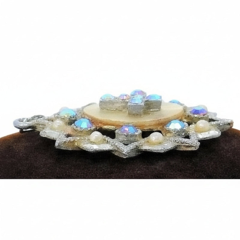 A vintage 1950s pendant featuring a cross with faceted Aurora Borealis stones and simulated pearls, set against an oval mother-of-pearl center, with intricate eloxal metalwork, profile.