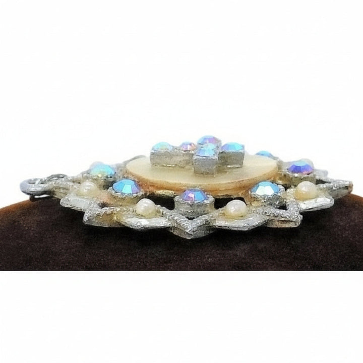 A vintage 1950s pendant featuring a cross with faceted Aurora Borealis stones and simulated pearls, set against an oval mother-of-pearl center, with intricate eloxal metalwork, profile.