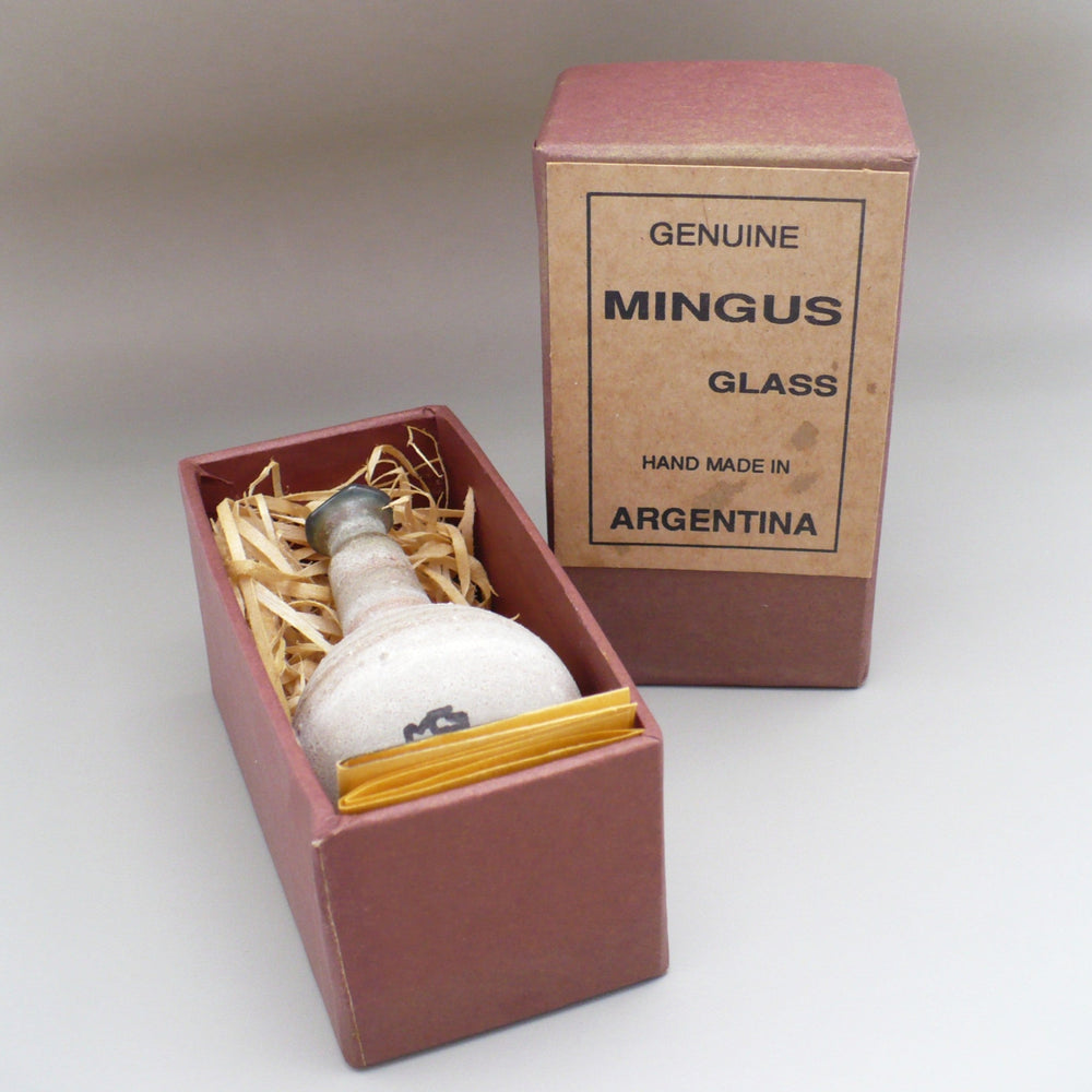 A vintage Mingus glass bottle presented in an open reddish-brown box with fine wood shavings, alongside its matching branded box on a neutral studio background.
