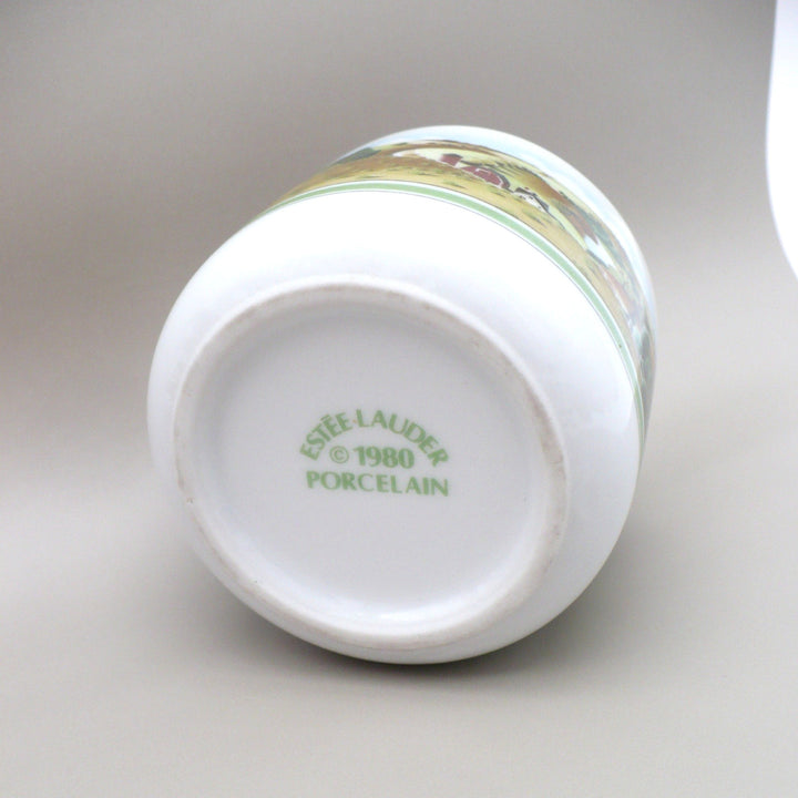 A white porcelain catch-all shown upside down, displaying the printed green text 'ESTEE LAUDER © 1980 PORCELAIN' on the bottom, with a decorative landscape band around the upper portion.