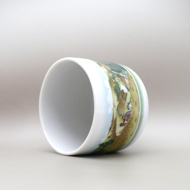 A white ceramic 1980 Estée Lauder Porcelain Trinket Catchall: American Folk Art Decor shown on its side, featuring a detailed landscape illustration on the side with green trees, a brown path, and a small red structure, photographed against a neutral gray background.