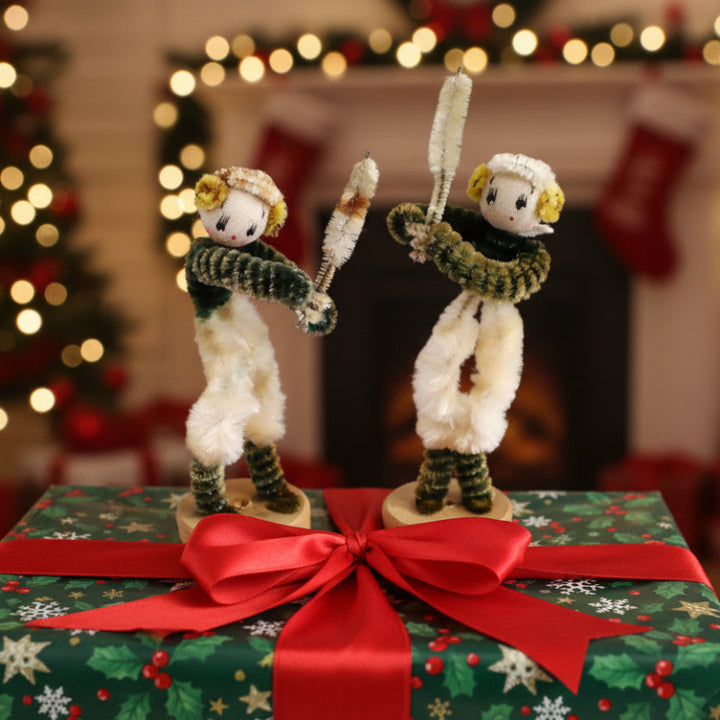 Two 1950s Japan Pipe Cleaner Baseball or Cricket Dolls – Vintage Sports Bendy Dolls. gift toppers on a Christmas-themed gift box with festive lights and stockings in the background.