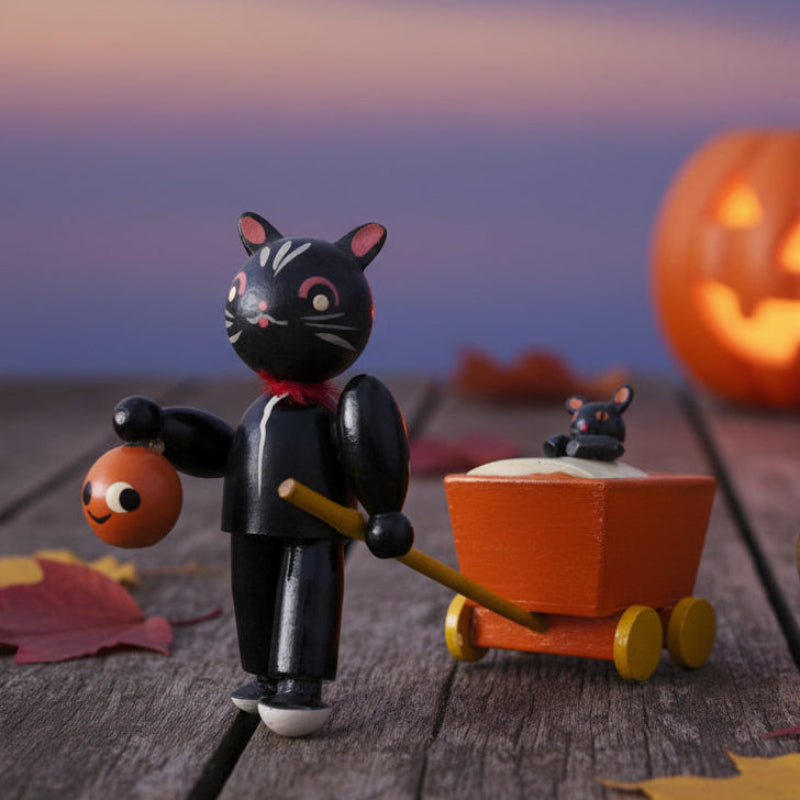 Black cat figurine with a mouse in a cart, pumpkin, and leaves on a wooden surface.