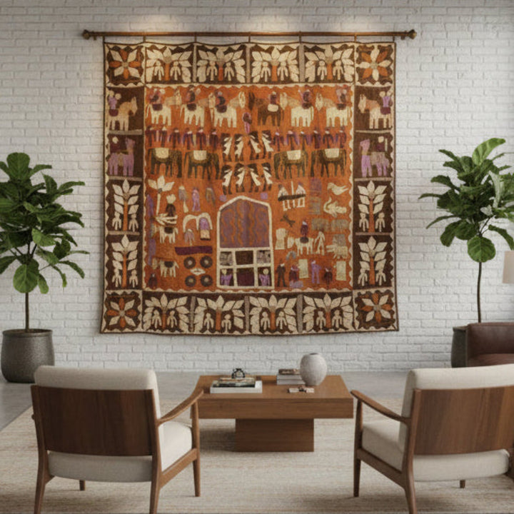 A20th-century Indian Kanduri shrine cloth featuring a detailed tribal-style scene with animals and geometric motifs, hanging on a white brick wall above a wooden coffee table and two armchairs in a modern living room setting.