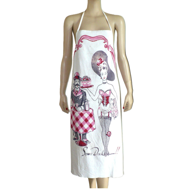 The image shows a white bib apron with a vintage-style print featuring a man and woman in a playful, suggestive pose. The man, dressed in a suit and hat, stands behind a table holding a dish of fruit and a drink, while the woman, in a burlesque-inspired outfit, stands beside him. The text "Some Dish!!" is printed at the bottom.