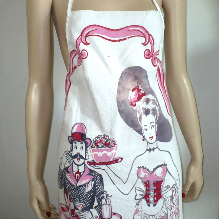 The image shows a white bib apron with a vintage-style print featuring a man and woman in a playful, suggestive pose. The man, dressed in a suit and hat, stands behind a table holding a dish of fruit and a drink, while the woman, in a burlesque-inspired outfit, stands beside him. The text "Some Dish!!" is printed at the bottom.