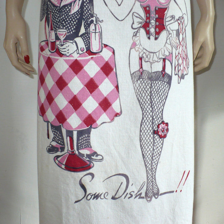 The image shows a white bib apron with a vintage-style print featuring a man and woman in a playful, suggestive pose. The man, dressed in a suit and hat, stands behind a table holding a dish of fruit and a drink, while the woman, in a burlesque-inspired outfit, stands beside him. The text "Some Dish!!" is printed at the bottom.