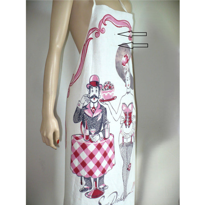 The image shows a white bib apron with a vintage-style print featuring a man and woman in a playful, suggestive pose. The man, dressed in a suit and hat, stands behind a table holding a dish of fruit and a drink, while the woman, in a burlesque-inspired outfit, stands beside him. The text "Some Dish!!" is printed at the bottom.