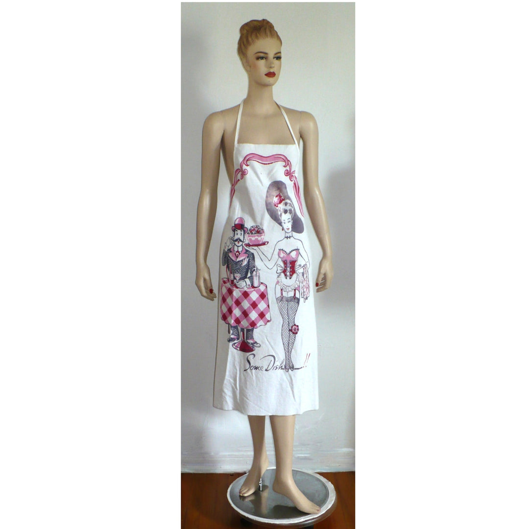 The image shows a white bib apron with a vintage-style print featuring a man and woman in a playful, suggestive pose. The man, dressed in a suit and hat, stands behind a table holding a dish of fruit and a drink, while the woman, in a burlesque-inspired outfit, stands beside him. The text "Some Dish!!" is printed at the bottom.