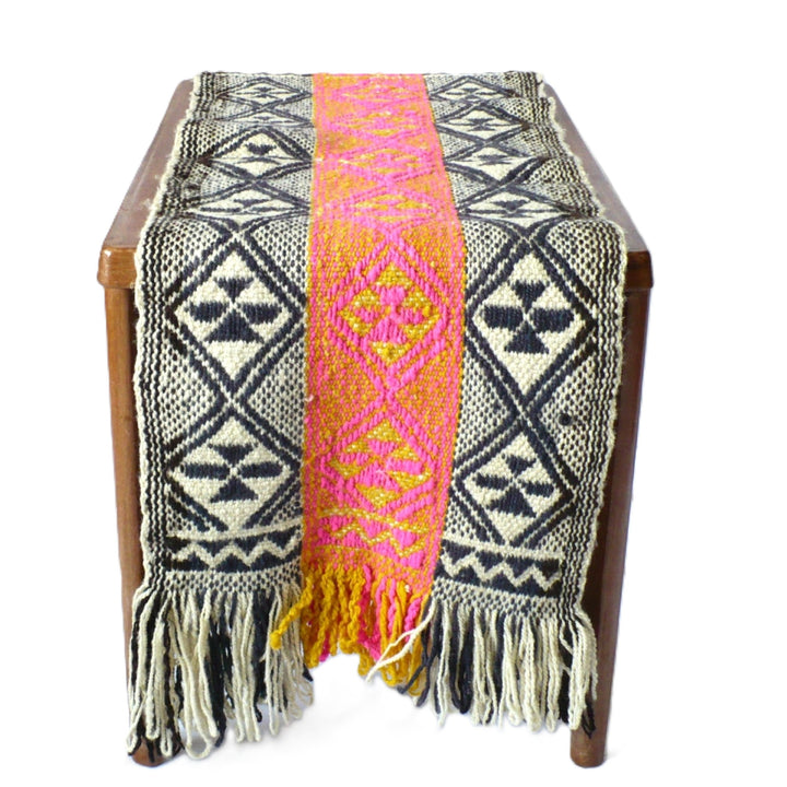 A 1960s Handwoven Mapuche Table Runner (42-inches) - Argentina, woven textile runner or throw draped over a wooden bench, featuring a bold geometric pattern in black and cream with a central vertical stripe in bright pink and yellow, finished with long looped fringe at the bottom.