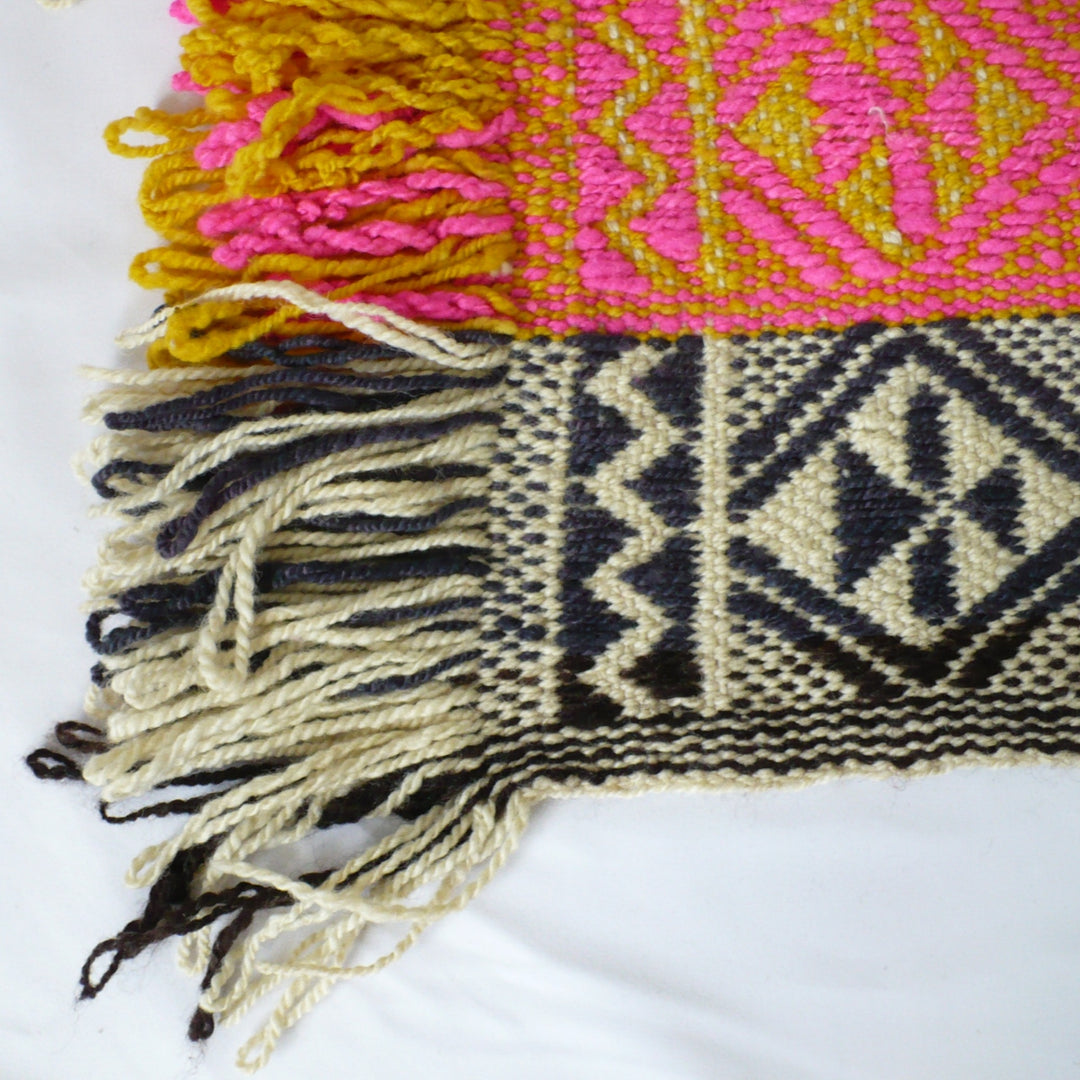 A close-up of a 1960s Handwoven Mapuche Table Runner, woven textile with fringe, showing a bright pink and mustard yellow patterned section, a cream and black geometric patterned section, and multicolored fringes on a white background.
