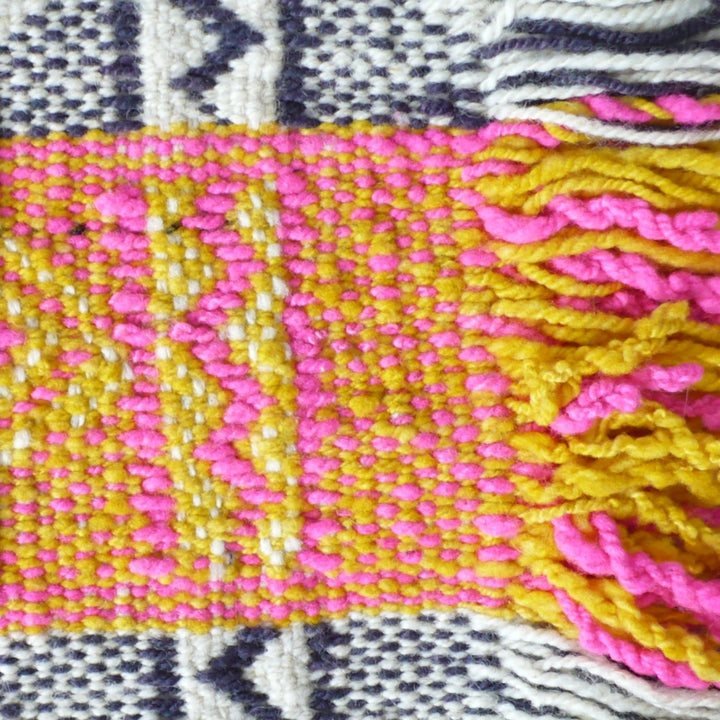 1960s Handwoven Mapuche Table Runner: Close-up of a woven wool textile with a bright pink and mustard yellow patterned section and pink-and-yellow fringe, bordered by blue and off-white woven sections.