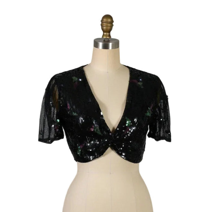 A black sequined sheer mesh bolero shrug with short puff sleeves and a deep V-neckline, displayed on a beige mannequin torso against a white background.