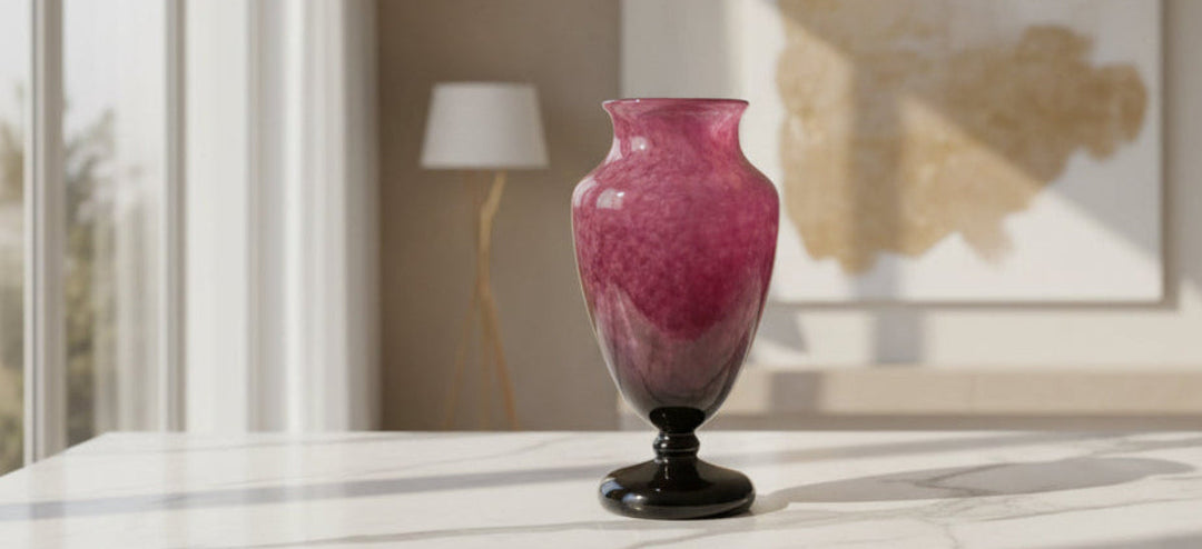 An antique vase with a textured, mottled pink and purple body and a glossy black pedestal base, in a beautifully lit room filled with natural sunlight.