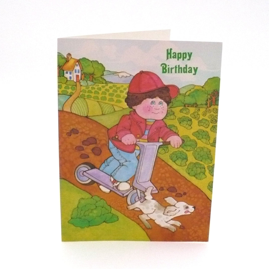 This authentic 1980s card features a classic Cabbage Patch Kids illustration by Current. The scene depicts a "Kid" in a red jacket and baseball cap riding a lavender scooter down a dirt path, accompanied by a playful, spotted puppy. In the background, the rolling green hills of the "Patch" are visible, dotted with cabbages and a small cottage.