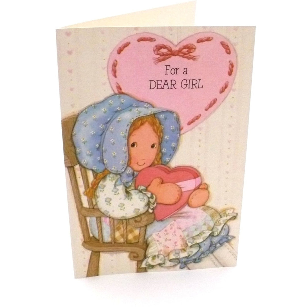 An illustrated vintage Valentine's Day card features a young girl in classic Holly Hobbie style, wearing a large blue-and-white floral sunbonnet and a patchwork dress. She is seated in a wooden chair, cradling a large pink heart-shaped box tied with a ribbon. Above her, a large pink heart contains the text "For a DEAR GIRL" against a cream-colored background with a subtle polka-dot and floral pattern.