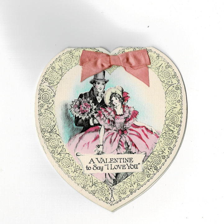 This elegant heart-shaped vintage Valentine’s Day card features a sophisticated construction where the sentiment is printed on a separate interior sleeve. The card follows a romantic Edwardian revival theme, with a real pink satin ribbon bow on the front that binds the interior sleeve to the die-cut outer shell.