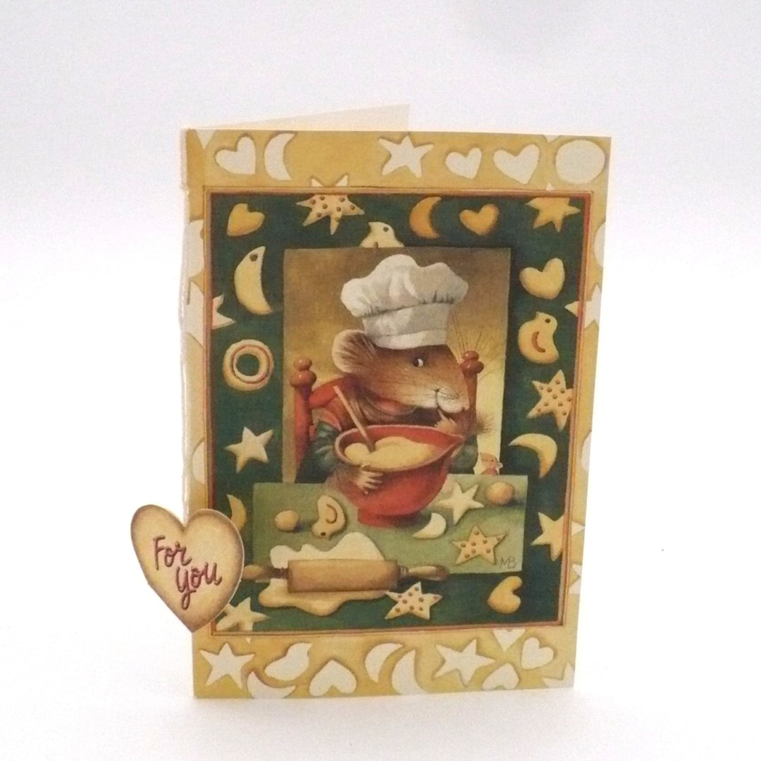 An illustration of an anthropomorphic mouse character named Vera the Mouse, dressed as a chef in a tall white toque and a red apron. She is shown in a cozy kitchen setting, diligently kneading or mixing dough on a floured wooden surface. The scene is framed by a decorative cream-colored border featuring a pattern of yellow crescent moons, stars, and hearts, giving it a whimsical, storybook feel. 
