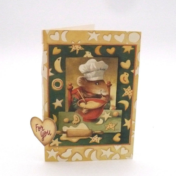 An illustration of an anthropomorphic mouse character named Vera the Mouse, dressed as a chef in a tall white toque and a red apron. She is shown in a cozy kitchen setting, diligently kneading or mixing dough on a floured wooden surface. The scene is framed by a decorative cream-colored border featuring a pattern of yellow crescent moons, stars, and hearts, giving it a whimsical, storybook feel. 