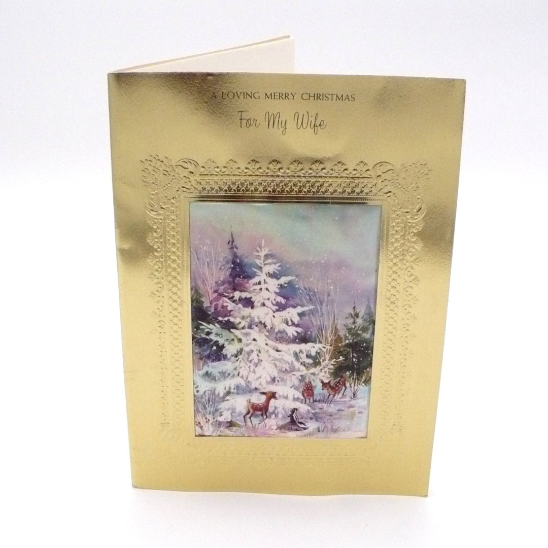 An upright vintage greeting card featuring a brilliant gold foil cover with an ornate, high-relief embossed frame. In the center is a rectangular die-cut window containing a raised, puffy fabric insert. The fabric is printed with a soft watercolor illustration of three brown deer gathered in a snowy forest around a large, snow-covered white spruce tree. Above the window, the text "A LOVING MERRY CHRISTMAS" is printed in an elegant script, followed by "For My Wife" below.