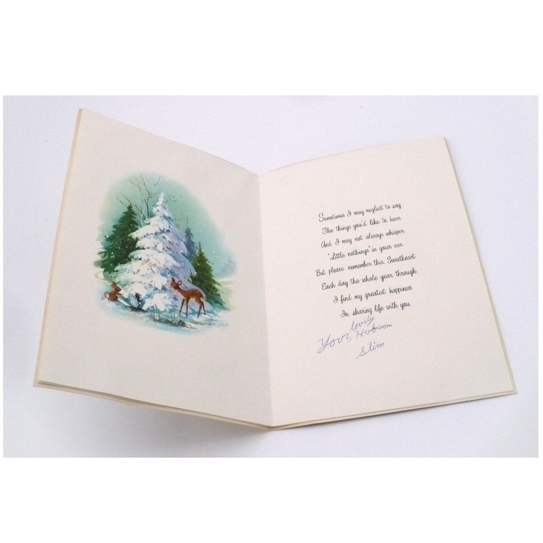 The open interior of a vintage greeting card made of heavy, cream-colored paper. On the left panel is a soft watercolor illustration of three brown deer gathered in a snowy clearing around a tall, snow-laden white spruce tree. On the right panel, a sentimental poem is printed in a dark, delicate calligraphic script. Below the printed text is a personal, hand-written signature in blue ink that reads, "Your loving husband, Slim."