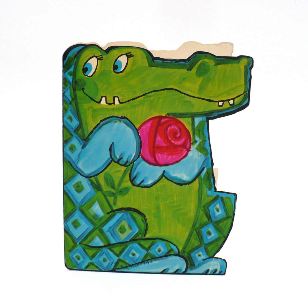 This whimsical greeting card features a hand-painted, friendly alligator depicted in a charming, folk-art style. The alligator is vibrant green with a playful blue-and-green diamond pattern on its side, and it looks toward the viewer with large, gentle blue eyes and a sweet, two-toothed smile. It holds a large, stylized pink rose close to its chest with soft blue paws, as if offering it as a gift. 