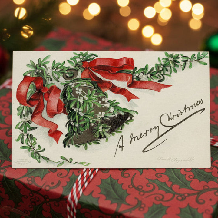 1907 Clapsaddle Christmas Bell & Mistletoe Vintage Postcard on a wrapped gift with festive lights and decorations.